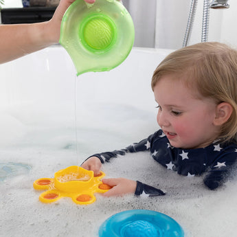 A child in a star swimsuit uses Fat Brain Toy Cos Dimpl Splash to enhance fine motor skills as water pours from a green container.