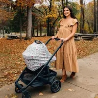 A woman in a brown dress pushes a stroller with the Milk Snob Winnie the Pooh Baby Bee Cover along an autumn tree-lined path.