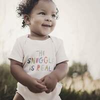 A happy baby in a Finn and Emma Organic Bodysuit, Snuggle Is Real, enjoys the sun amid lush green grass.
