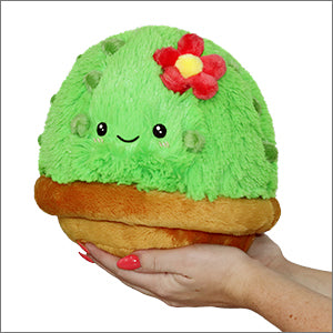 Hands holding a Plush, Mini Cactus by Squishable—a cuddly toy with a red flower and smile, crafted from soft polyester fiber.