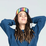 A smiling person with dreadlocks wears a blue sweater and Storied Hats Adult Hat, Pride, hands on head against a light gray background.
