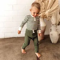 A toddler in a Snuggle Hunny striped shirt and Organic Pants, Olive, walks barefoot on concrete, framed by a white brick wall and vases.