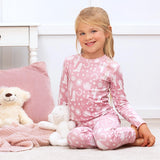 A girl in Tesa Babes Wild West Pink PJ set sits on the floor, surrounded by stuffed animals, a pink pillow, and a knit blanket.