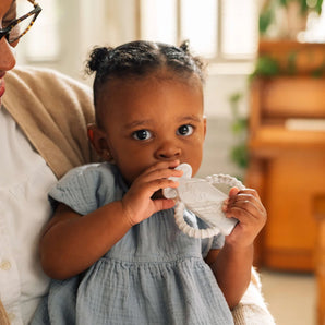 A baby with pigtails holds a Milk Snob Calm n Cuddle™ Cookies & Cream teether while wearing a light blue dress on an adults lap.