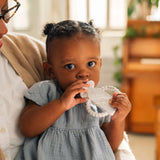 A baby in a blue dress holds a Milk Snob Calm n Cuddle™ Teether Cookies & Cream, sitting on an adults lap indoors.