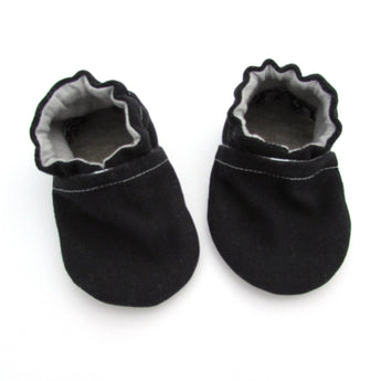 Cabooties Denim Black: Small black baby shoes with white stitching, ruffled edges, and flexible rubber soles on a white background.
