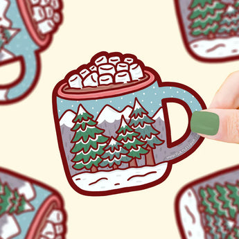 A hand with green nails holds a Turtle Soup Vinyl Sticker mug filled with hot cocoa, featuring mountains and trees.