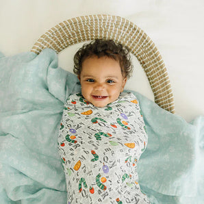 A smiling baby wrapped in a vibrant LouLou Lollipop Muslin Swaddle Set, Eric Carle, rests on a light blue sheet with a wicker pillow.