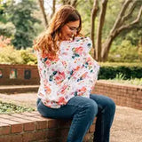 A woman breastfeeds outdoors under a Milk Snob Cover, wearing jeans and a matching French Floral top. She has brown hair and glasses.