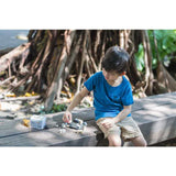 Young boy in a blue shirt plays with a PlanToys Construction Set on a wooden bench, surrounded by large tree roots.