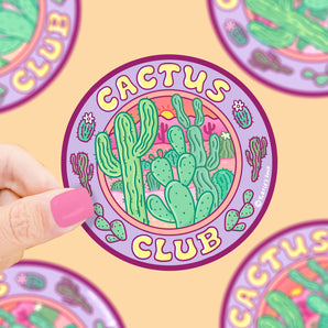 A hand with pink nails holds a Cactus Club vinyl sticker by Turtle Soup, featuring green cacti on a vibrant purple background.