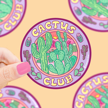 A hand with pink nails holds a Cactus Club vinyl sticker by Turtle Soup, featuring green cacti on a vibrant purple background.