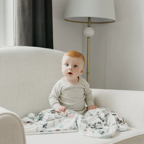 A baby with red hair sits on a white armchair, wrapped in Copper Pearls Jo Knit Swaddle Blanket, with a floor lamp behind.