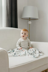 A baby with red hair sits on a white armchair, wrapped in Copper Pearls Jo Knit Swaddle Blanket, with a floor lamp behind.
