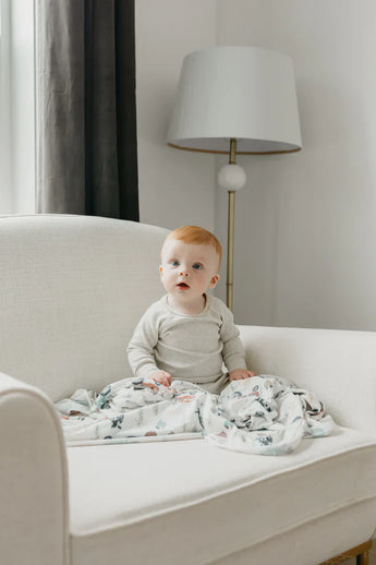 A baby with red hair sits on a white armchair, wrapped in Copper Pearls Jo Knit Swaddle Blanket, with a floor lamp behind.