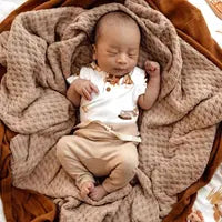 A sleeping baby in a white shirt and Snuggle Hunnys Organic Pants, Pebble lies on a soft, textured brown blanket.