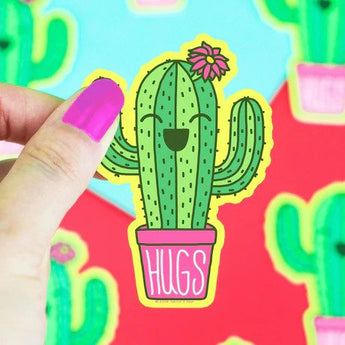 A hand with purple nails holds a Turtle Soup Vinyl Sticker of a smiling cactus in a pink pot labeled Hugs with a pink flower.