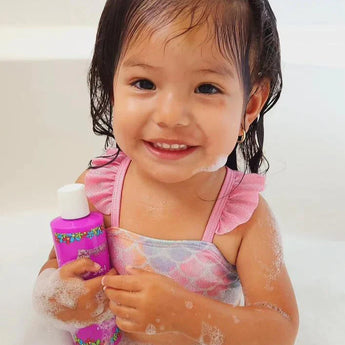 A smiling child in a bubble bath holds a pink Piggy Paint 3-in-1 Shampoo, Body Wash, Bubble Bath with wet hair and a swimsuit.