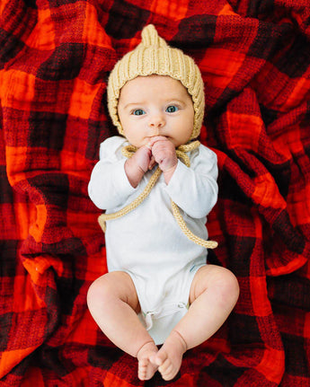 A baby with wide eyes lies under a Little Unicorn Cotton Swaddle, Red Plaid, wearing a knitted hat and white outfit.