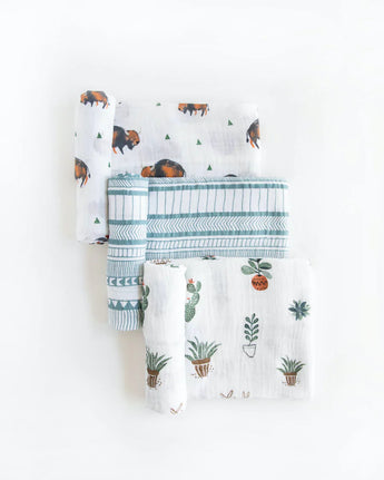 Set of three folded muslin swaddles by Little Unicorn with bison, geometric lines, and potted plants on white.