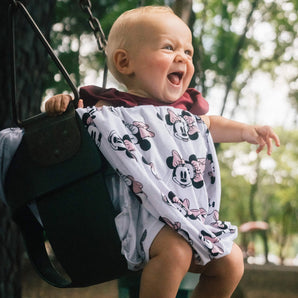 A baby in Cover Disneys Minnie Mouse by Milk Snob laughs joyfully while sitting on a swing at the park.