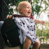 A baby in Cover Disneys Minnie Mouse by Milk Snob laughs joyfully while sitting on a swing at the park.