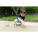 A child crouches to play PlanToys Meadow Ring Toss, aiming at pegs on concrete, honing fine motor skills with grass behind.