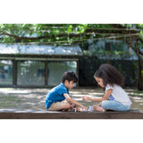 Two kids enjoy their PlanToys Construction Set on a wooden platform, surrounded by greenery and trees.