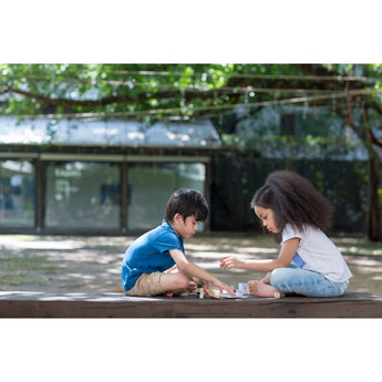 Two kids enjoy their PlanToys Construction Set on a wooden platform, surrounded by greenery and trees.