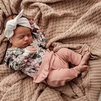 A baby sleeps on a beige blanket in a floral top, Snuggle Hunnys Rose organic pants, and a white bow headband.