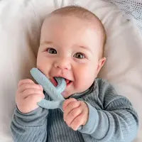 Baby in a blue onesie smiles, holding Ryan & Roses non-toxic Cactus Teether Toy in Mist for soothing relief on a white cushion.