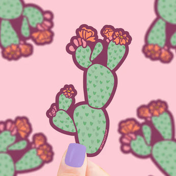 Hand holds Turtle Soups Vinyl Sticker, Prickly Pear Cactus, with pink flowers on a light pink cactus-patterned background.