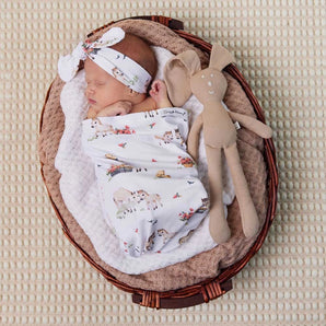 A newborn snoozes in a Snuggle Hunny Organic Snuggle Swaddle & Topknot Set, Pony Pals, nestled with a stuffed bunny.