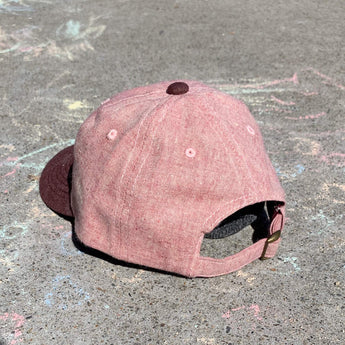 The Lil Hat Jam by Storied Hats, a sustainable pink cap from UV-resistant hemp, lies on a pavement with colorful chalk drawings.