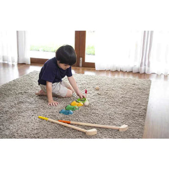 Child playing on a carpet with colorful blocks near PlanToys Mini Golf Set, wearing a navy shirt and beige shorts.