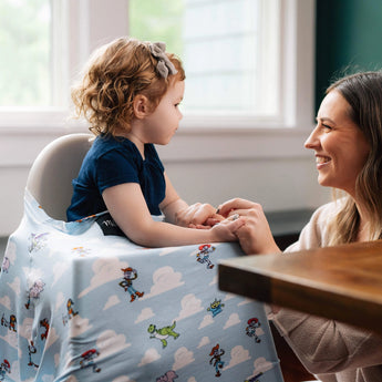 A woman smiles at a toddler on a high chair with a Milk Snob Cover, featuring Disney & Pixars Toy Story design for multifunctional charm.