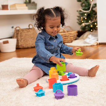 A toddler in a denim dress hones fine motor skills with the Plugzy Shape Learning Toy by Fat Brain Toy Co, amid decor of trees and baskets.