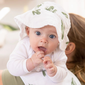 A curious blue-eyed baby in Bebe Au Laits Saguaro Sun Hat lies on an adults shoulder.