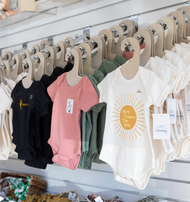 A display of baby onesies hangs on a white wall rack in a store. The onesies are in various colors, including black, pink, and white, with different designs and text. Shelves with additional baby products are partially visible below.