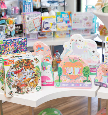 A store display featuring various colorful puzzle boxes. The designs include fairy tales, animals, and princess themes. The display is on white tables in a well-lit shop, with more toys visible in the background.