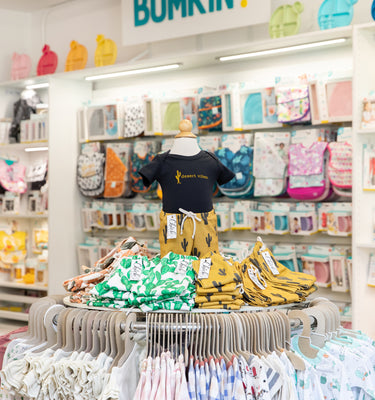 A childrens clothing store with colorful displays. The foreground has a round rack with various patterned outfits, topped with a mannequin wearing a black top. Shelves in the background display bags and more clothing items.