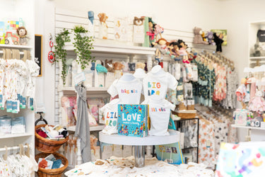 Display of childrens clothing and gifts in a store. Mannequins show BIG BRO, LIL SIS tees. Shelves are stocked with toys, books, and clothes. A woven basket and plush toys add to the cozy atmosphere.