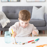 In a bright room with a gray sofa, a child paints at a white table while wearing a Bumkins Long-Sleeved Art Smock: Wander.