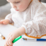 A child wears a Bumkins Long-Sleeved Art Smock: Wander, painting with a green paintbrush at a white table, brushes scattered nearby.