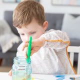 A child wearing a Bumkins Long-Sleeved Art Smock: Wander places a paintbrush into a jar, focused on creativity at a white table.