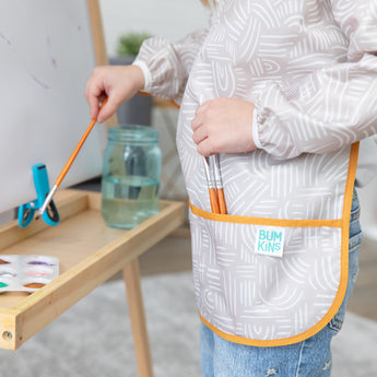 A child paints at an easel in a Bumkins Long-Sleeved Art Smock: Wander, with paints and water in a jar nearby.