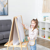A girl with a bow in her hair paints at an easel, wearing a Bumkins Long-Sleeved Art Smock: Wander, in a cozy living room.
