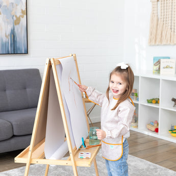 A girl with a bow in her hair paints at an easel, wearing a Bumkins Long-Sleeved Art Smock: Wander, in a cozy living room.
