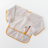 Bumkins Long-Sleeved Art Smock: Wander lays on a white background, holding two paintbrushes in the front pocket.