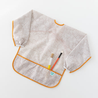 Bumkins Long-Sleeved Art Smock: Wander lays on a white background, holding two paintbrushes in the front pocket.